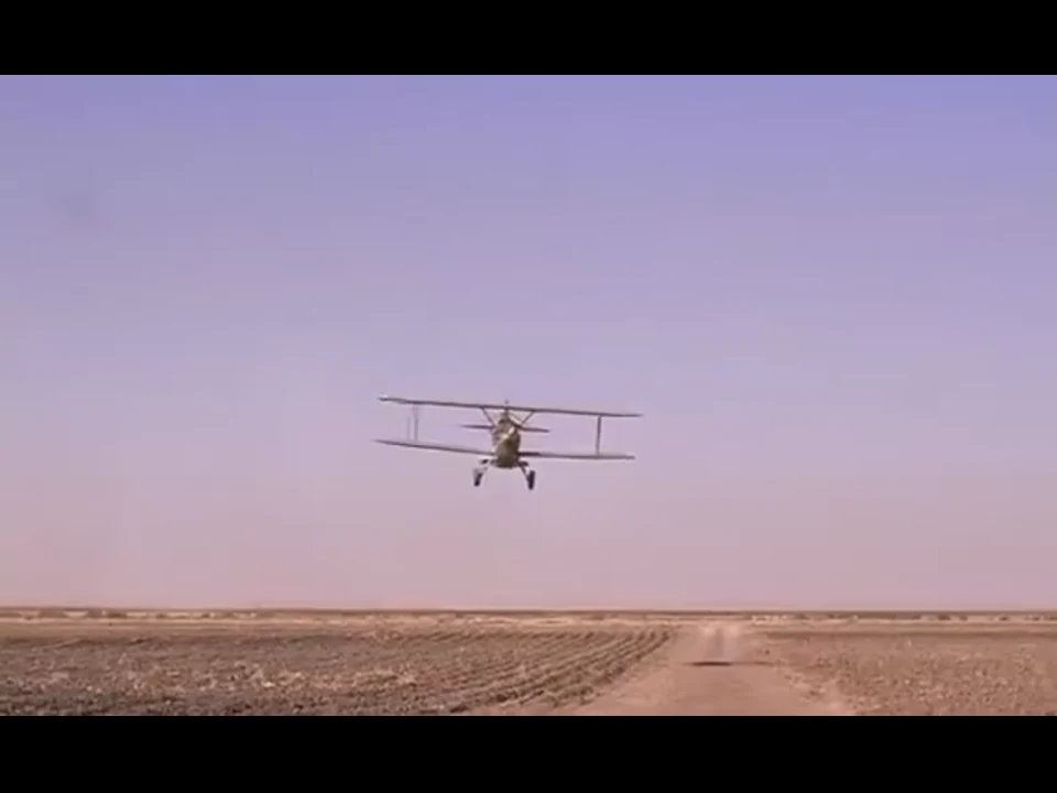 一架双翼飞机正面低飞，机身压近荒地与土路的地平线。