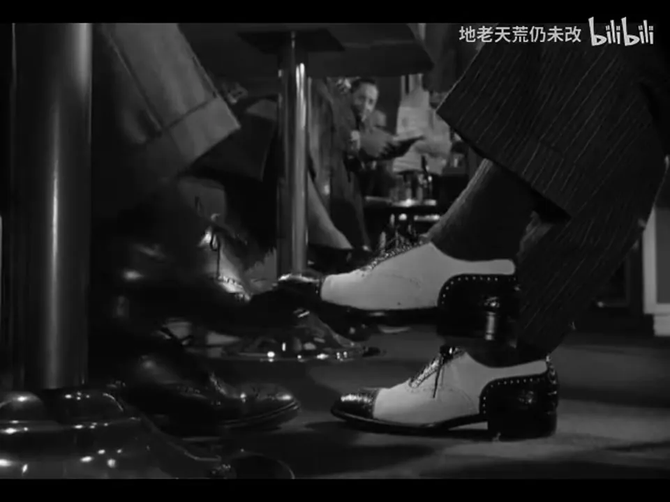 火车包厢座位下,Guy 从左侧移来的深色鞋碰到 Bruno 右侧的双色便鞋;Bruno 另一只双色鞋停在右下方,画面没有显示二人的脸。