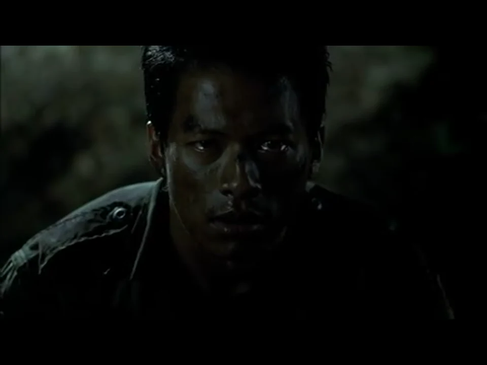 士兵满脸汗水与泥污，在黑暗中正面凝视画外，眼睛被微光照亮