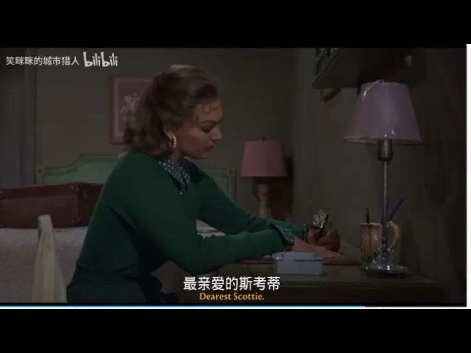 Judy 身穿绿色上衣坐在旅馆房间书桌前写信;画面底部的英文硬字幕可读为“Dearest Scottie”。
